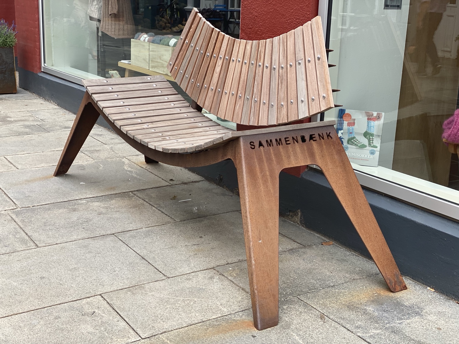 Public bench with curved wooden slats on a corten steel frame, its seat dipping gently toward the middle