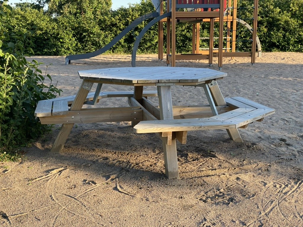 Round wooden picnic table in a sandy playground with gaps cut between the attached bench sections. A climbing frame and slide behind.