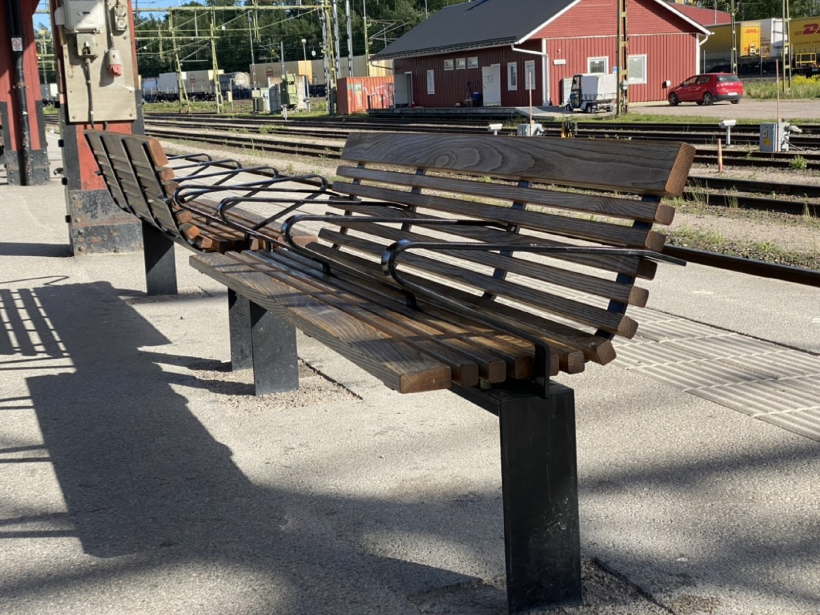 Two identical wooden-slatted benches on a Swedish rail platform, mirrored so that each faces its own track. Each bench is flat, with a slatted backrest and four black steel armrests that curve out from the backrest and anchor in the seat.