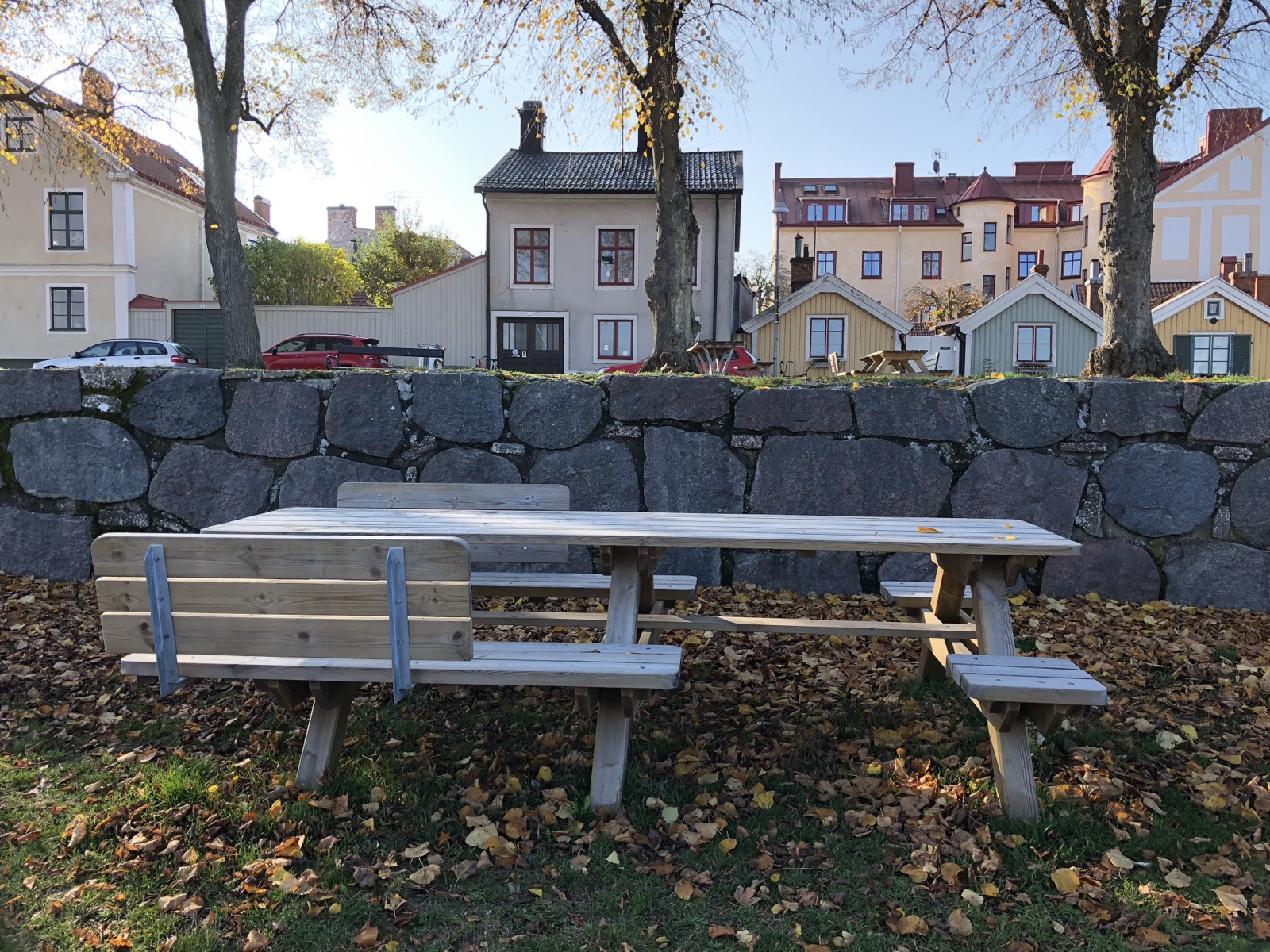 Long wooden picnic table on leaf-covered grass below Kalmar's old stone city wall. The attached seating varies along the table, bench sections with backrests and sections without, and two open cutouts in the seating line face each other near the middle.