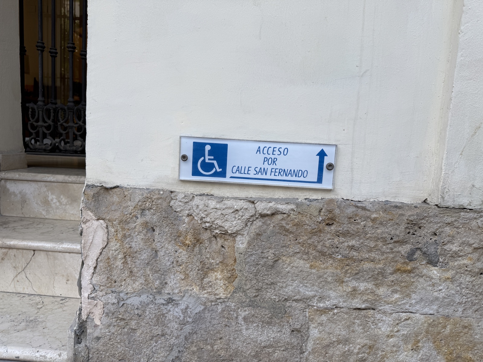 Small white plaque on a worn stone wall in Alicante, a blue wheelchair symbol and the text Acceso por Calle San Fernando with an arrow turning up and round the corner. To the left, marble steps lead up to a metal-grille door.