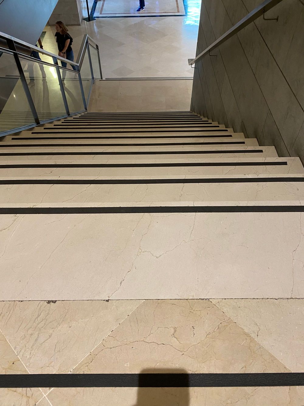 A marble staircase seen from the top, each step edged with a dark contrast strip, with a glass railing on the left and a handrail on the right.