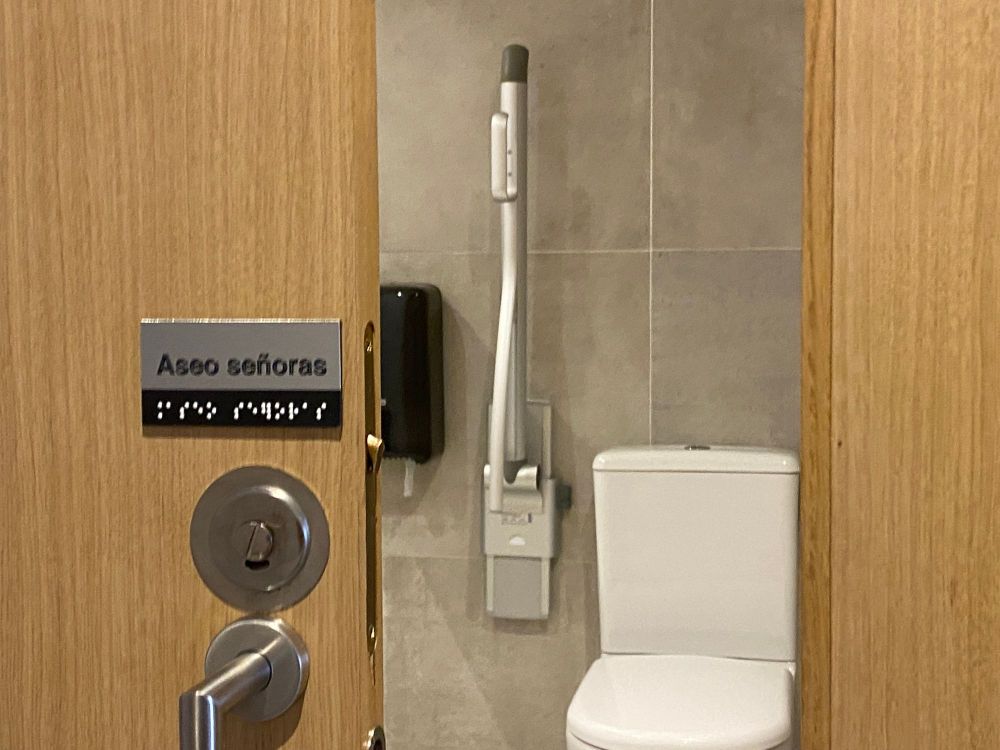 A wooden door with the sign "Aseo señoras" and braille beneath it. Through the open door, a toilet with a folded-up support arm beside it.