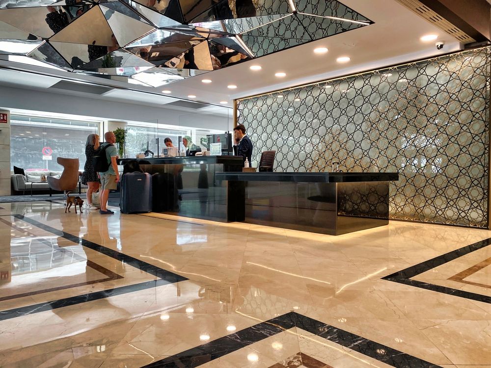 The same reception seen from the right. The dark counter steps down to a lower section nearest the camera. Guests with luggage and a small dog stand at the far end, and the mirrored ceiling reflects the lobby.