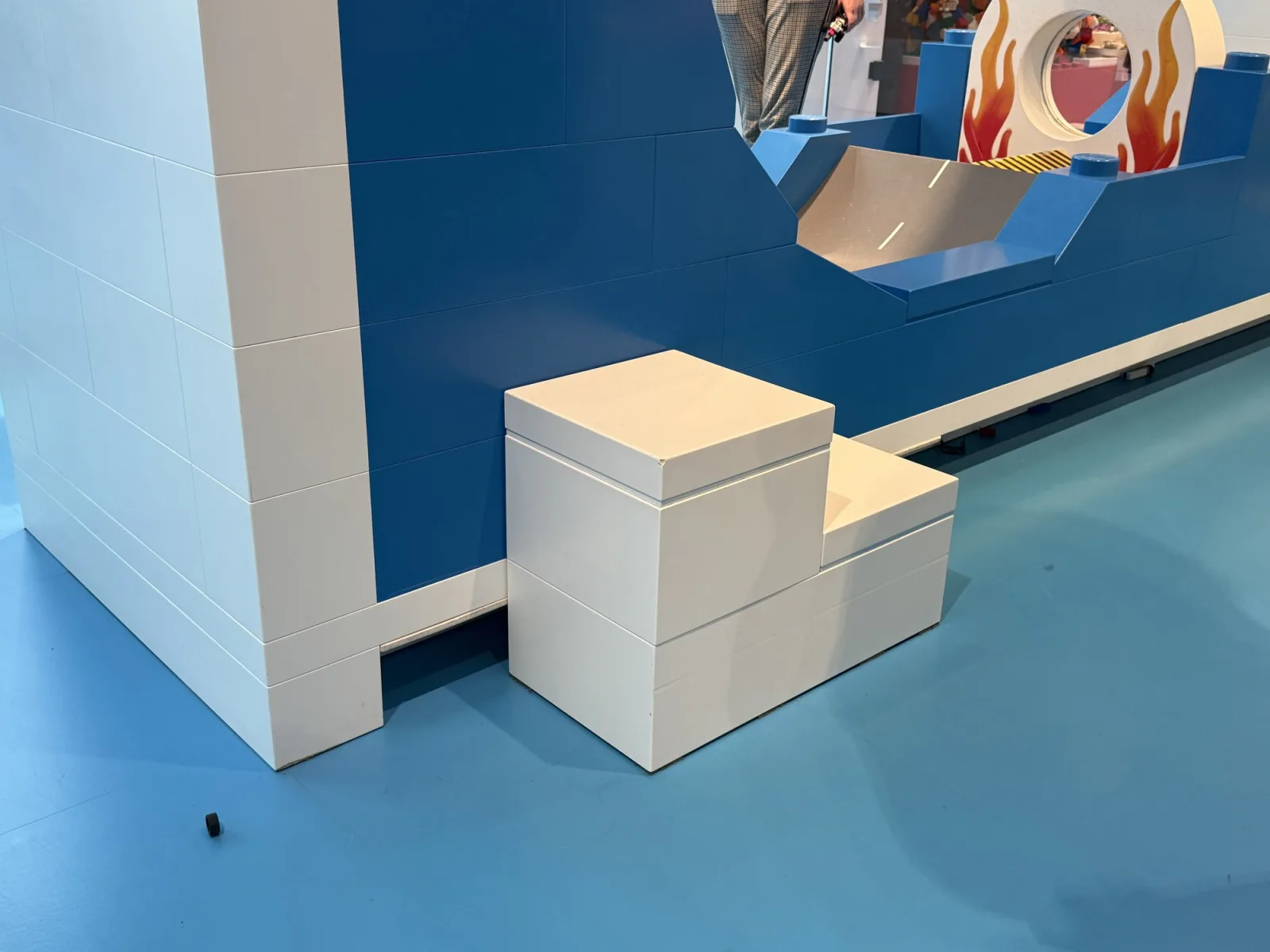 A loose white step with two heights in one piece stands on a blue floor, next to a blue and white wall built like large LEGO bricks.