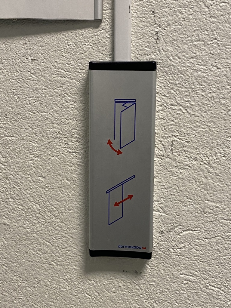 A grey wall-mounted button with two blue line drawings: a door swinging open with a red arrow, and a door sliding sideways with a double red arrow.